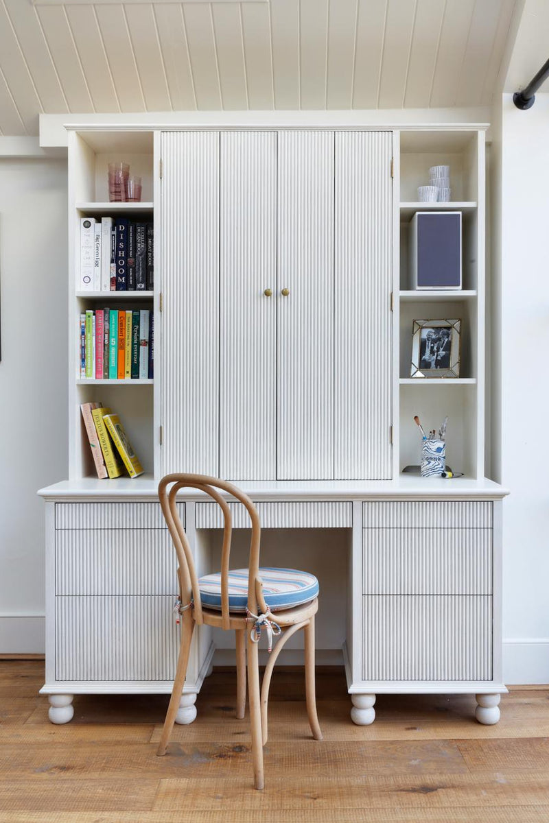 A white built-in desk and cabinet unit with open shelving, reeded cabinetry and turned ball feet.
